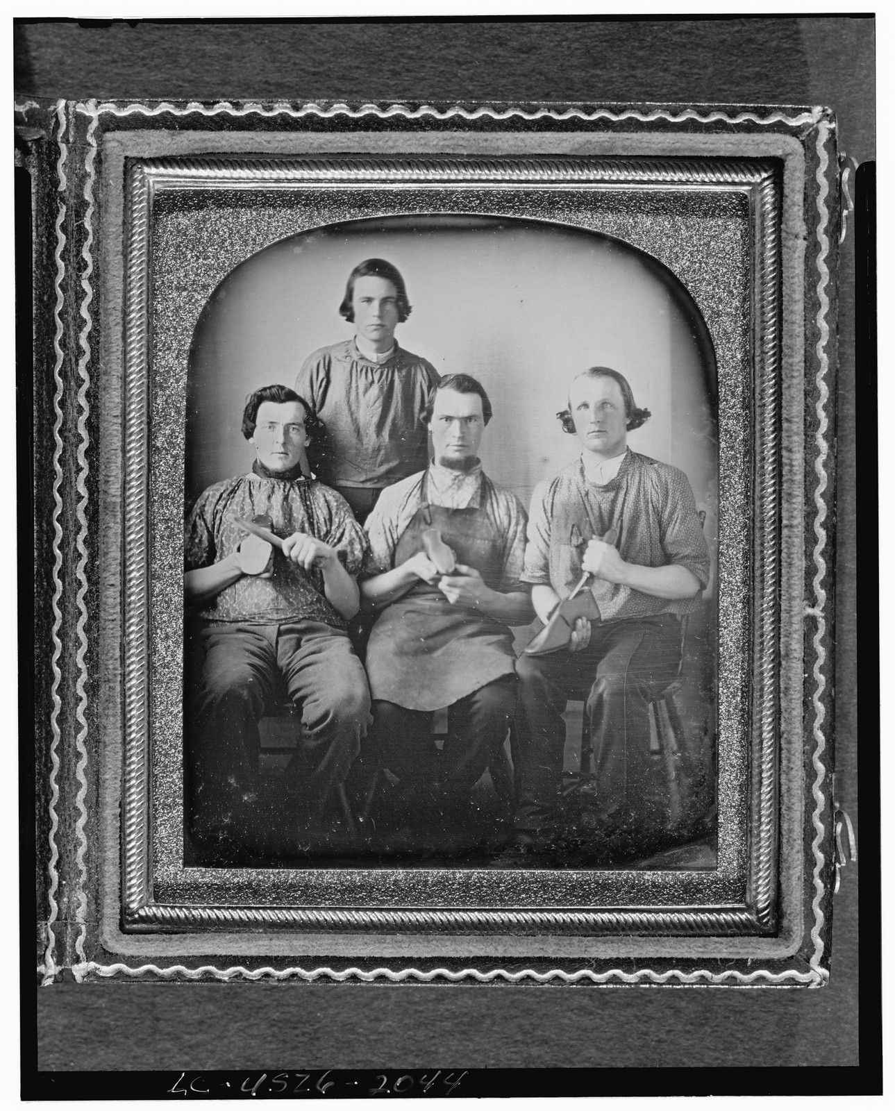 [Occupational group portrait of four shoemakers, one full-length, standing, other three seated, holding shoes and shoe making equipment], [between 1840 and 1860] — historic photograph