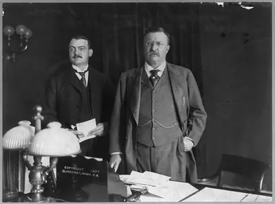 [Theodore Roosevelt, Pres. U.S., half length, standing behind desk, facing slightly left, with his asst. secy. William Loeb]