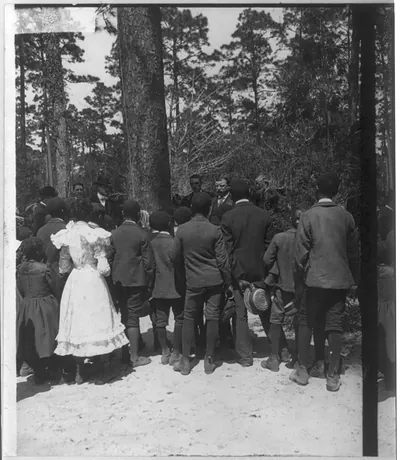 [Theodore Roosevelt, bust, standing in front of wooded area, facing left, speaking to group consisting of mainly African American children]
