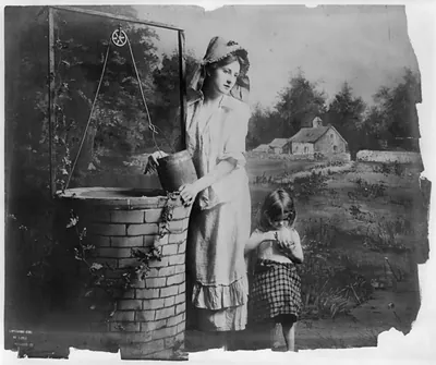 [Young woman modeling: Full lgth., standing at well, with little girl]