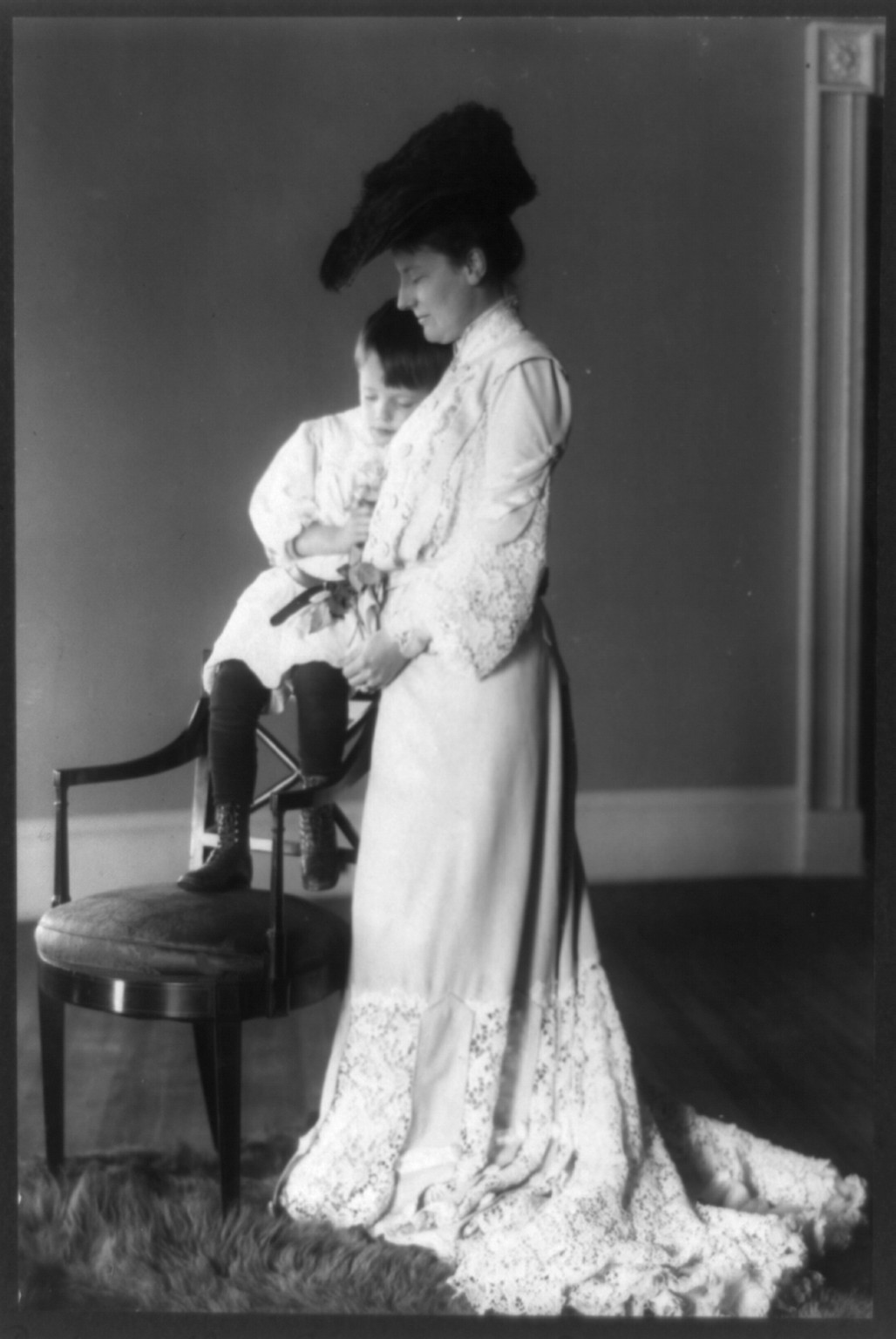 [Edith Kermit Carow Roosevelt, full-length portrait, standing, facing left, with arm around young Quentin Roosevelt, who is seated on back of chair], c1903. — historic photograph