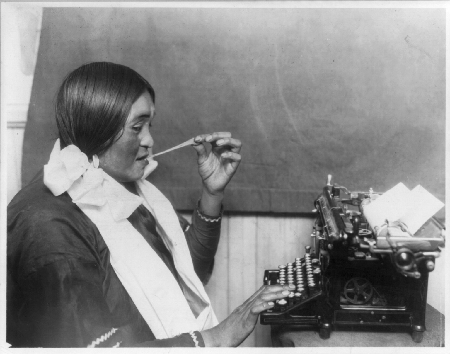 [Mrs. Many Horse typing for the Indian agent on the Glacier National Park Reservation and stretching chewing gum from her mouth], [no date recorded on caption card] — historic photograph
