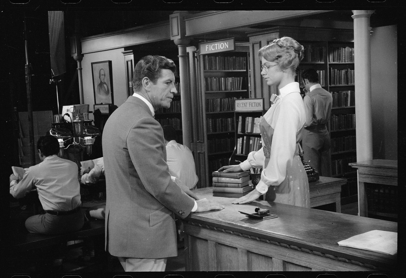 Man and woman at a library desk beside Fiction shelves — historic image