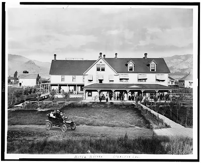 [Hotel Sierra, Loyalton, California, with people standing on porch, people in automobile in foreground, and mountains in background]