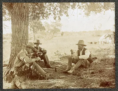 Cowboys sitting under a tree, one eating from a tin