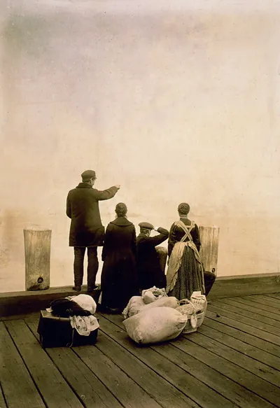 [Four immigrants and their belongings, on a dock, looking out over the water; view from behind]