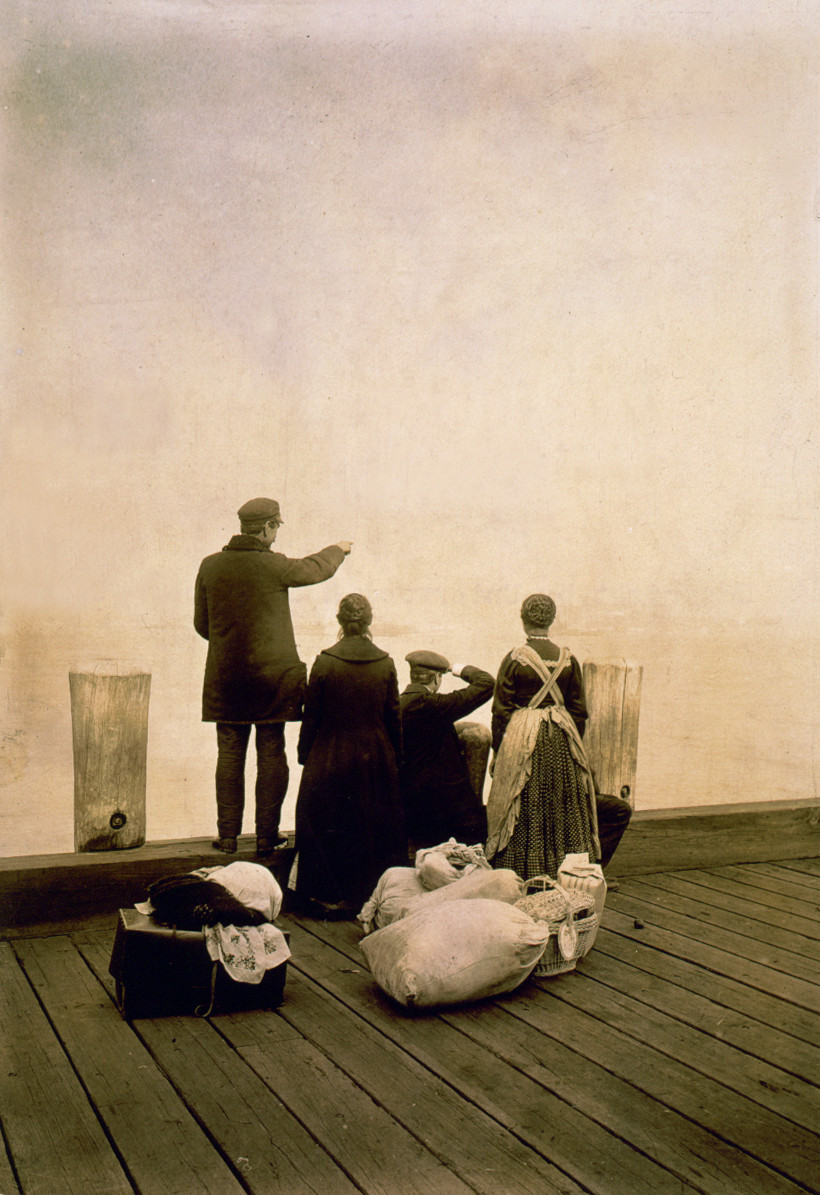 [Four immigrants and their belongings, on a dock, looking out over the water; view from behind], c1912 Oct. 30. — historic photograph