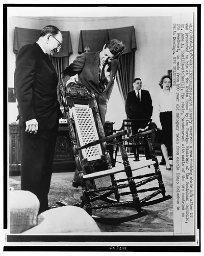 [President John F. Kennedy examines mahogany rocking chair given by Dominican Republic, as Foreign Minister of the Dominican Republic Dr. José A. Bonilla Atiles (l) looks on]