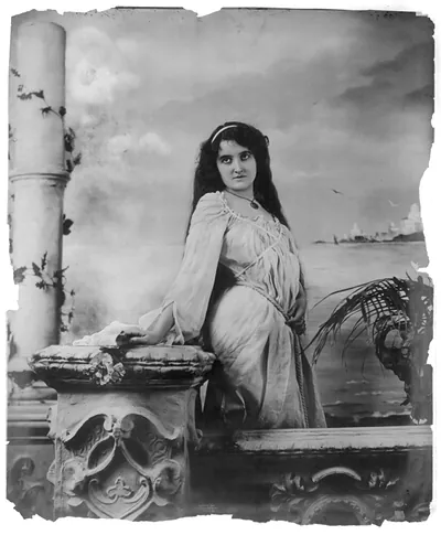 [Young woman modeling: 3/4 lgth., standing by wall, in front of body of water]