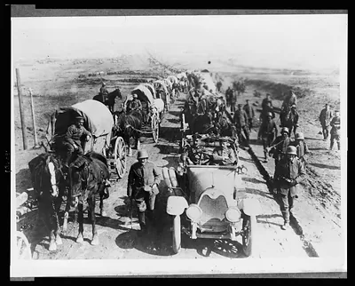 [German officers in an automobile on the road with a convoy of wagons; soldiers walk along side the road]