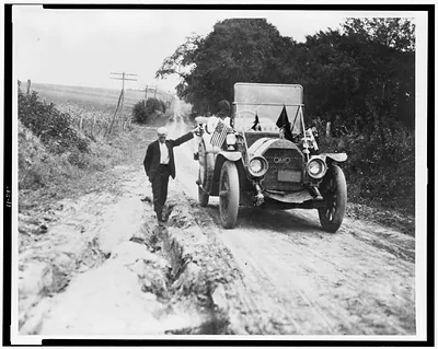 [Man standing in rut, with hand on automobile]