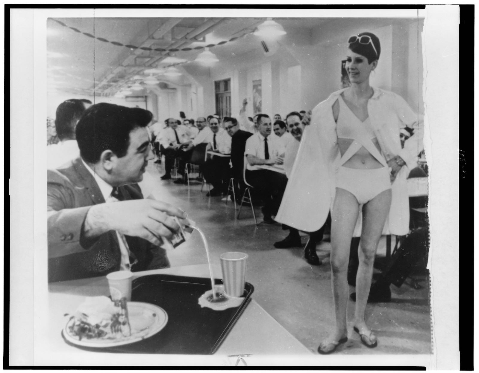 [Vince Lagrotteria looking at Karin Fester, who is modeling a bathing suit, in the cafeteria of the Pratt & Whitney machine tool plant in West Hartford, Conn.] — historic photograph