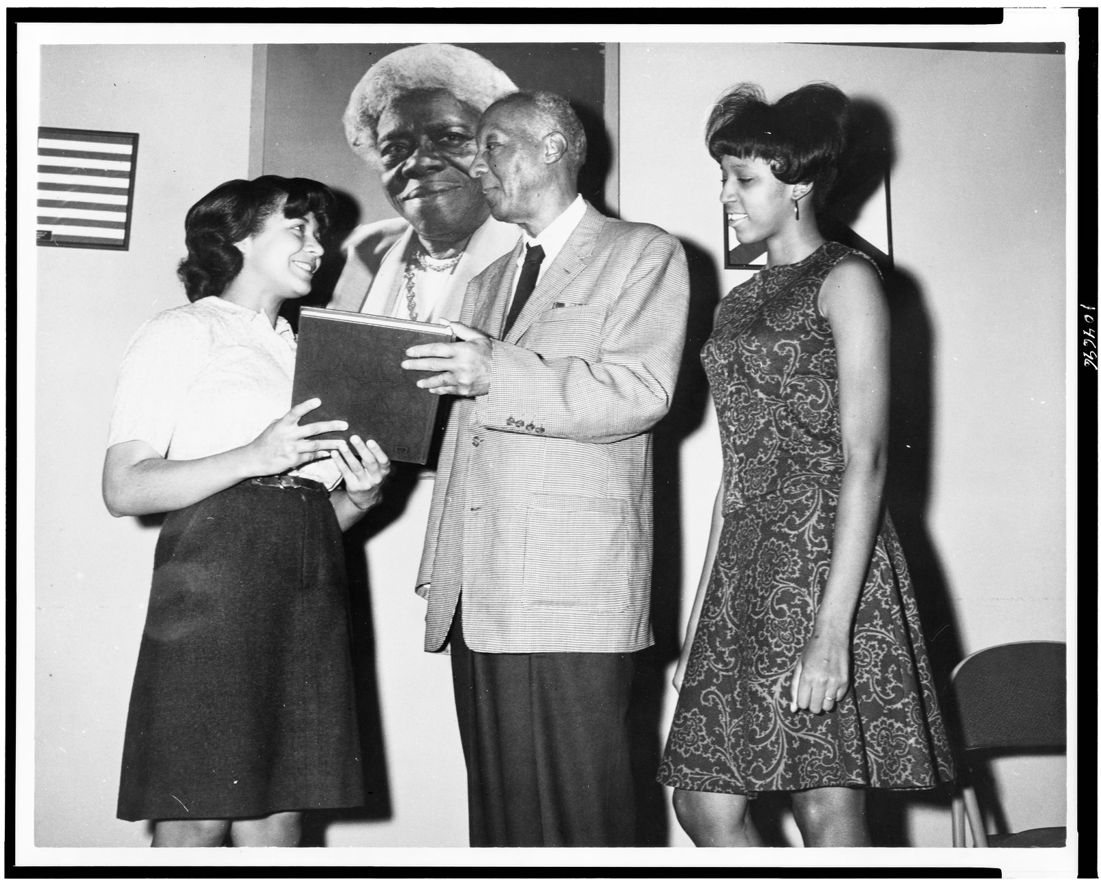 Miss Joyce Hanks, senior, Bethune-Cookman College, Crescent City, Florida, presents A. Phillip Randolph, also of Crescent City, Florida, a copy of the yearbook with Mary Finley, sophomore of Ocala, Florida, looking on. They stand in front of the last professional picture taken of Mrs. Mary McLeod Bethune, founder of the college / photographed by Martin Gross., [1967 Apr.] — historic photograph