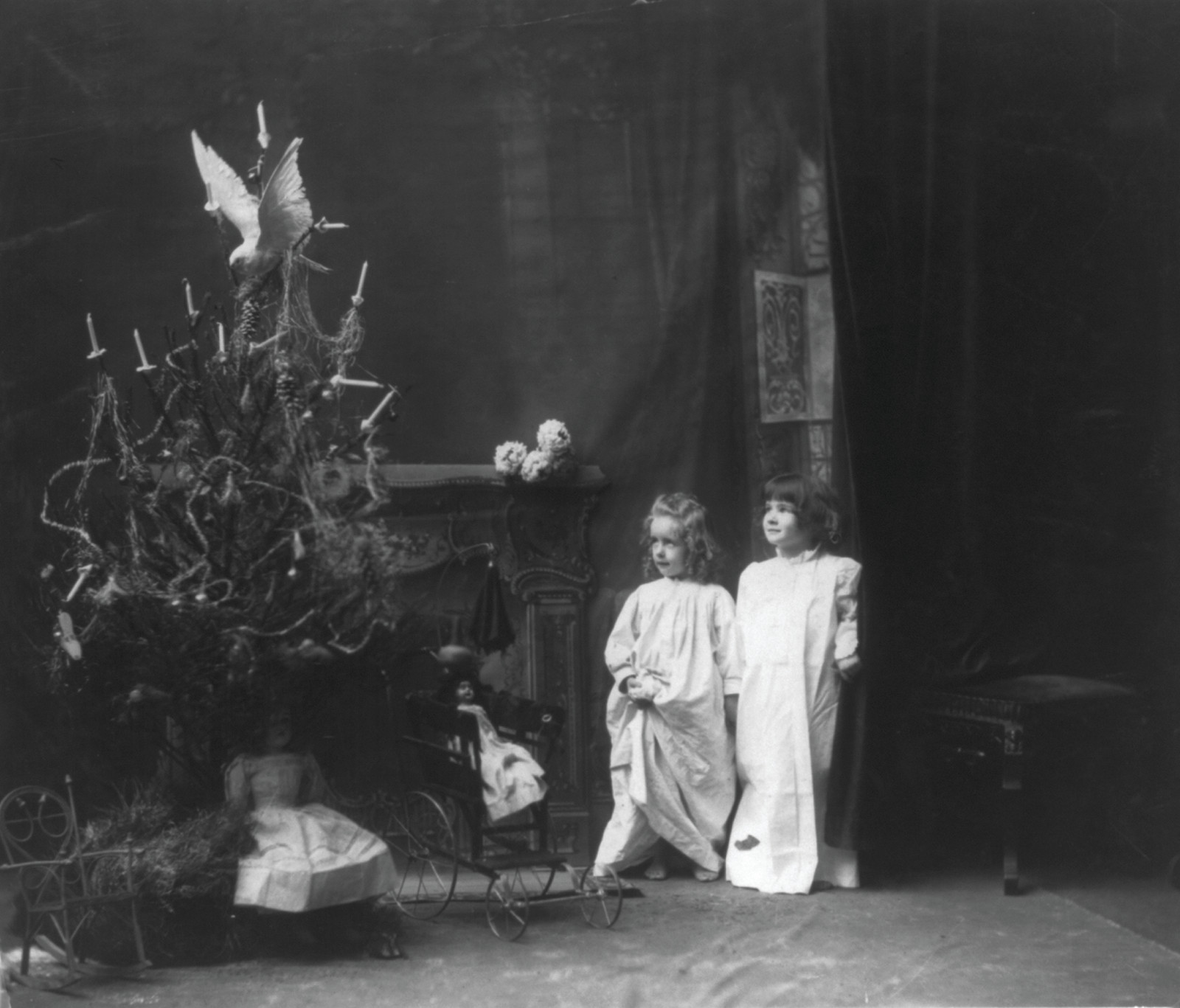 [Sentimental Christmas scenes of children and trees: 2 small girls in nightgowns looking at decorated tree; dolls, chair and carriage] — historic photograph