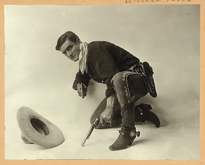Cowboy in studio pose kneeling with two revolvers beside a hat