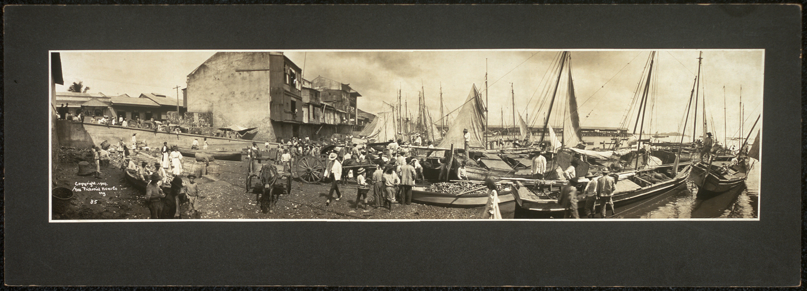 Panoramic sepia photograph of a waterfront with sailing vessels and crowds — historic photograph