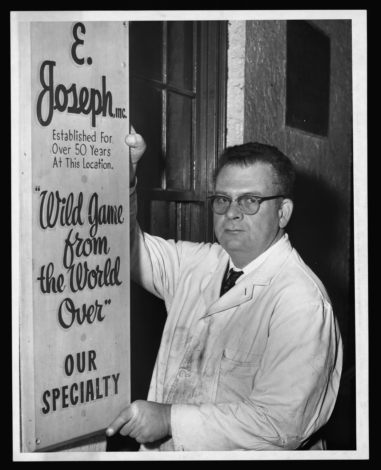 Man in white coat beside E. Joseph, Inc. sign — historic image
