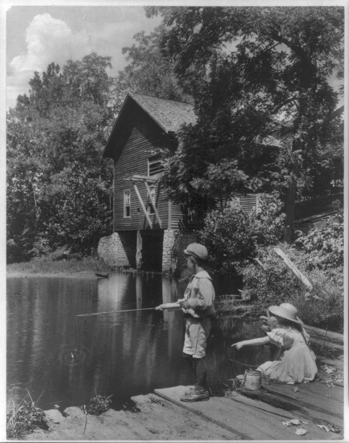 [Rural scenes of children fishing: boy and girl at pond behind barn] — historic photograph