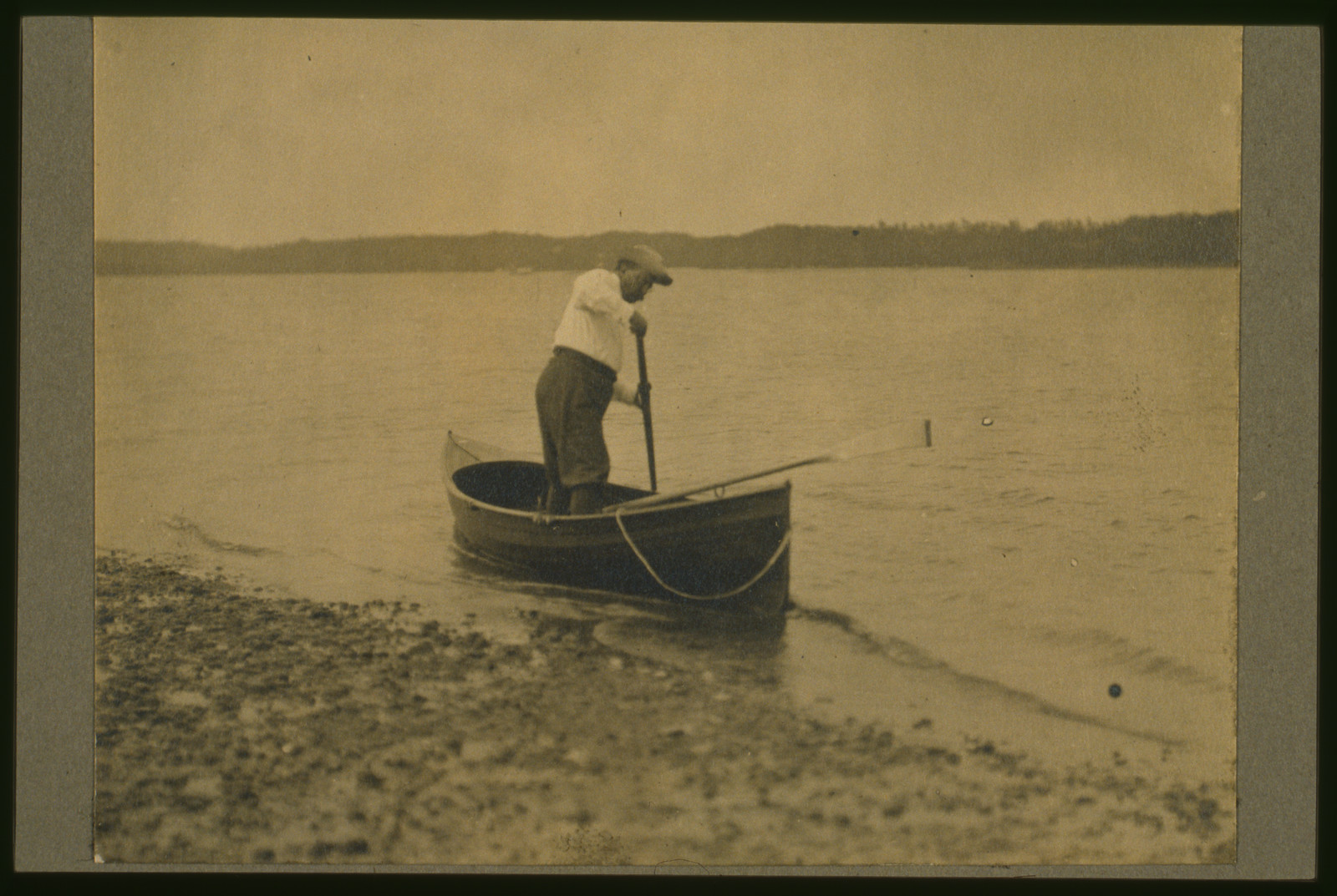 [Theodore Roosevelt, standing up in rowboat, pushing away from shore with oar], c1905. — historic photograph