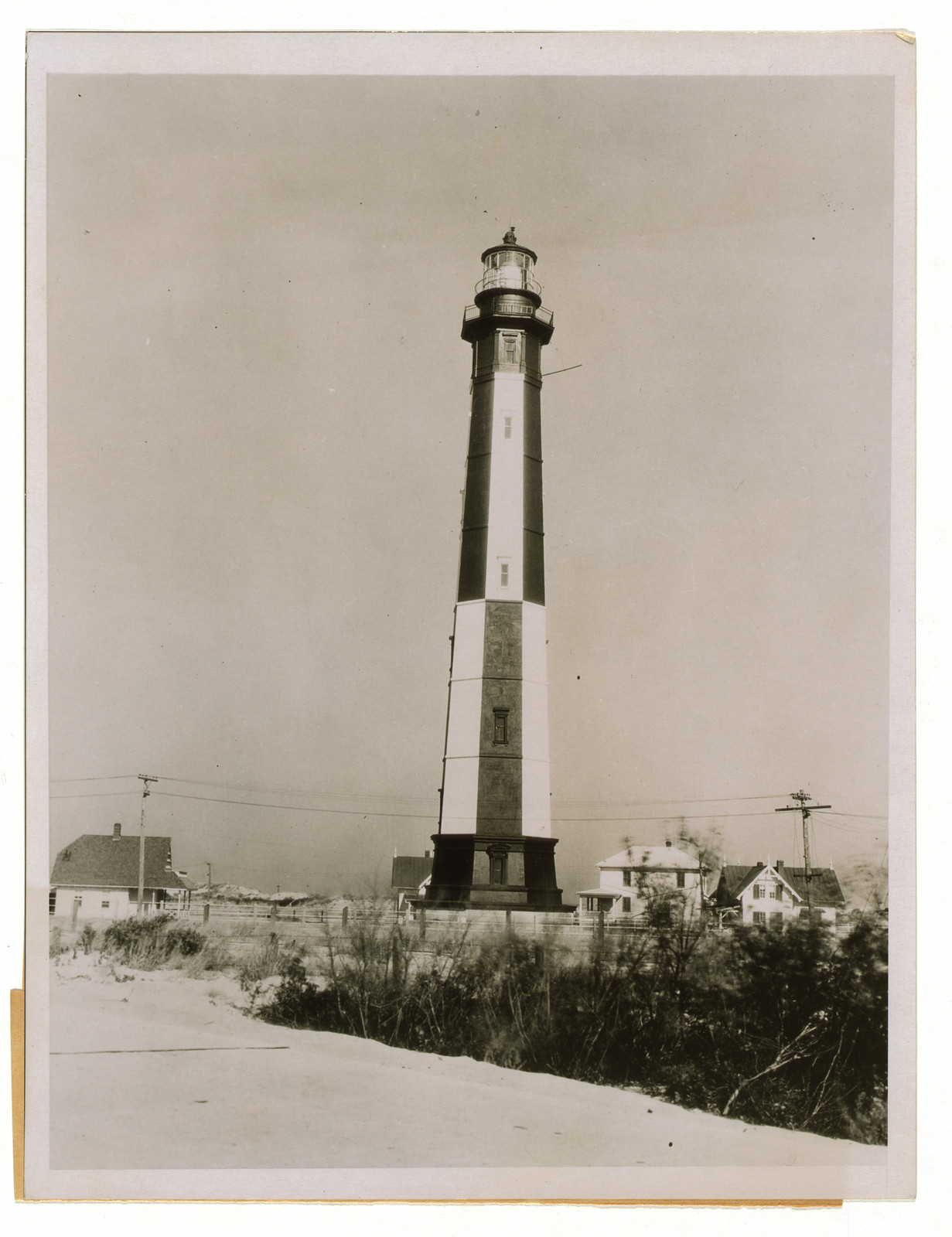 Black and white striped lighthouse with dunes and houses — historic image