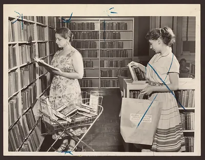 Two women choosing books from library shelves with a book cart