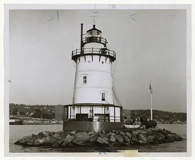 Lighthouse on rocky island in a harbor, black-and-white photograph