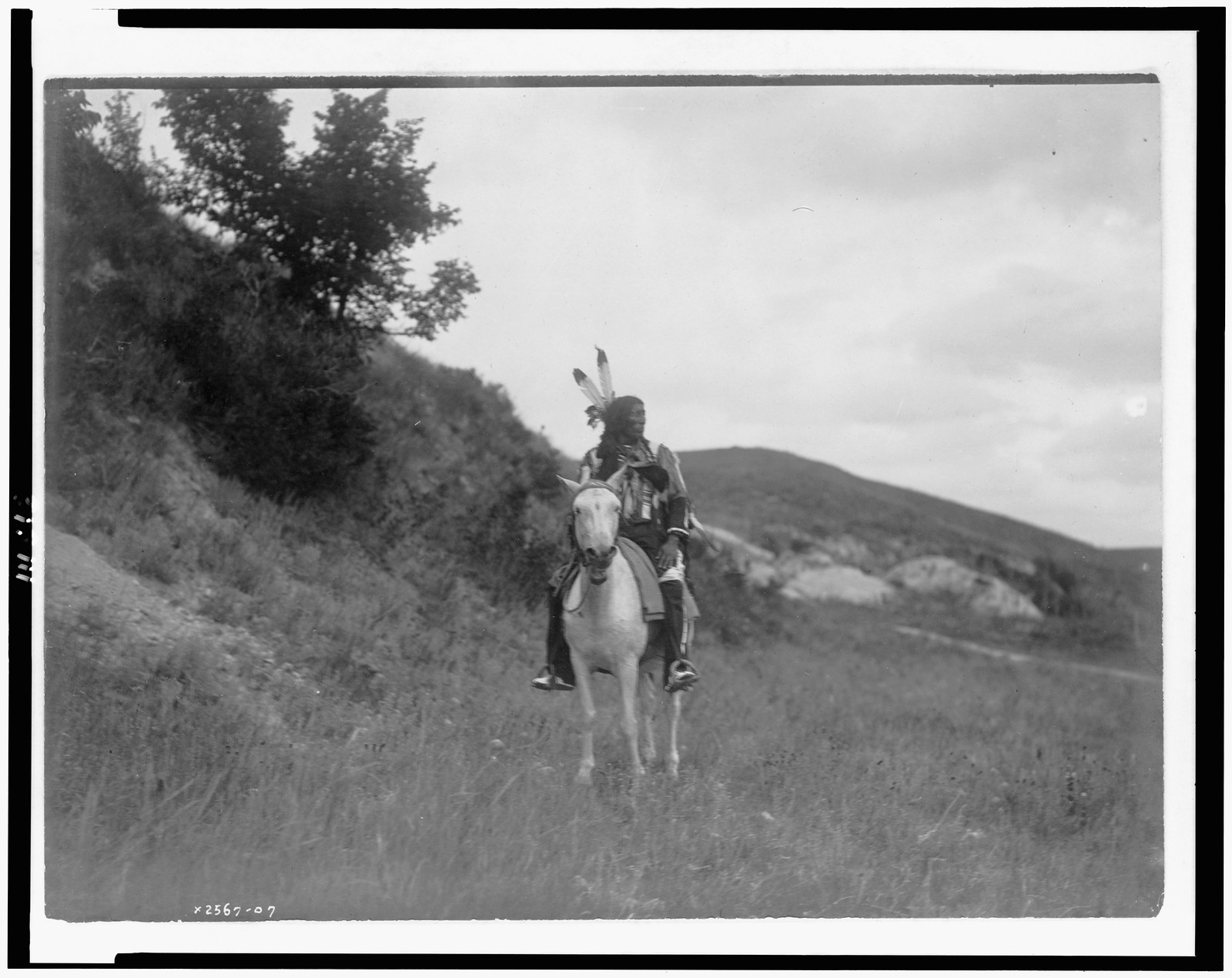 [Sioux Indian on horseback, wearing two feathers, beaded buckskin shirt, and leggings, with hills in background], c1907. — historic photograph