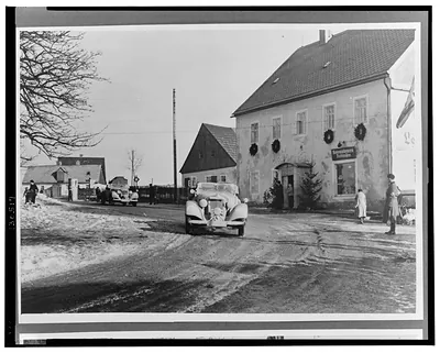 [Two Mercedes-Benz automobiles passing in front of a building decorated with wreaths]