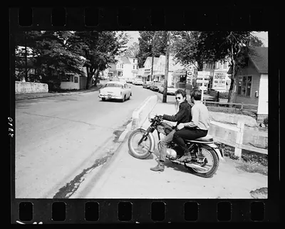 [Bob Dylan riding on motorcycle in street, with another man seated behind him] / Gilbert-Look.