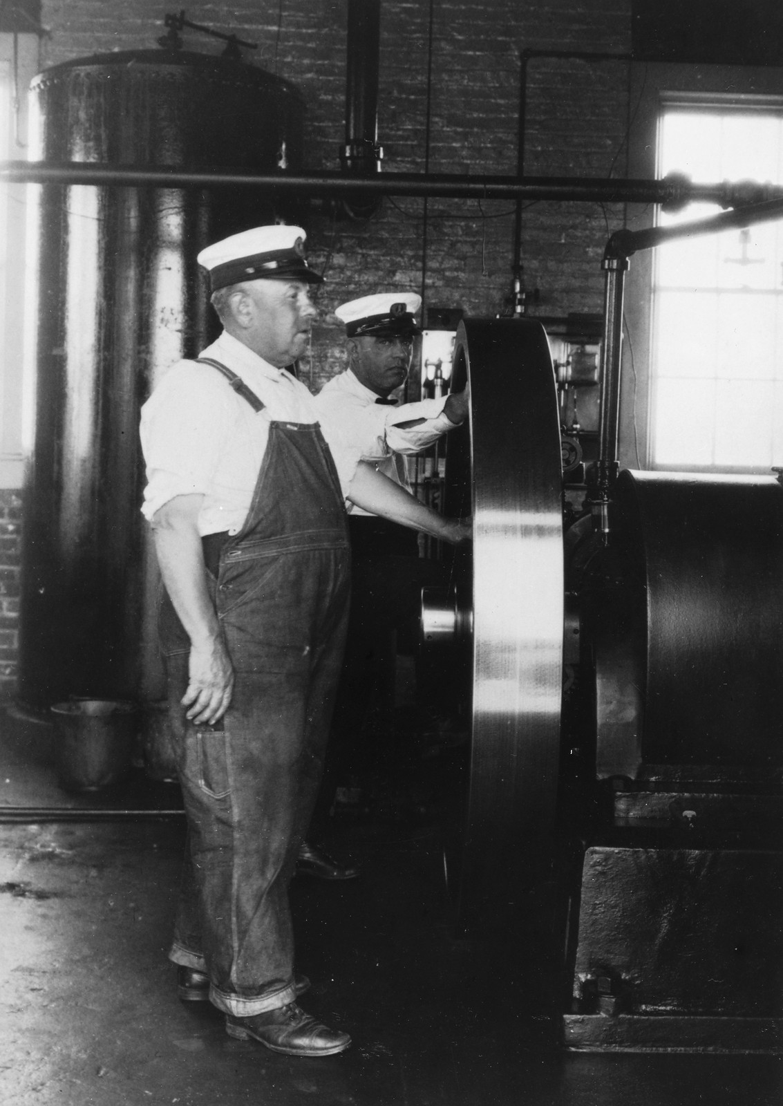 Two men in caps beside a large engine flywheel — historic image