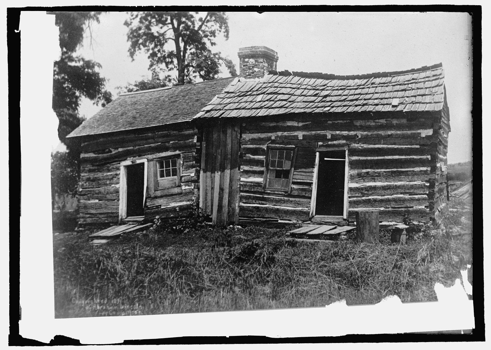 Log cabin with stone chimney and shingled roof — historic image