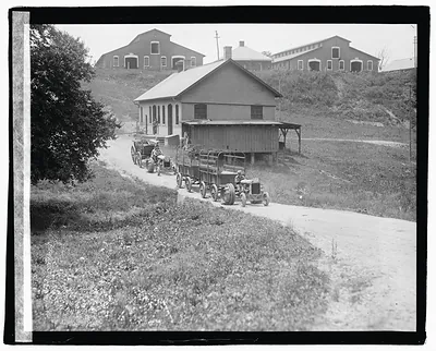 Two tractors pulling wagons on a farm road, barns behind