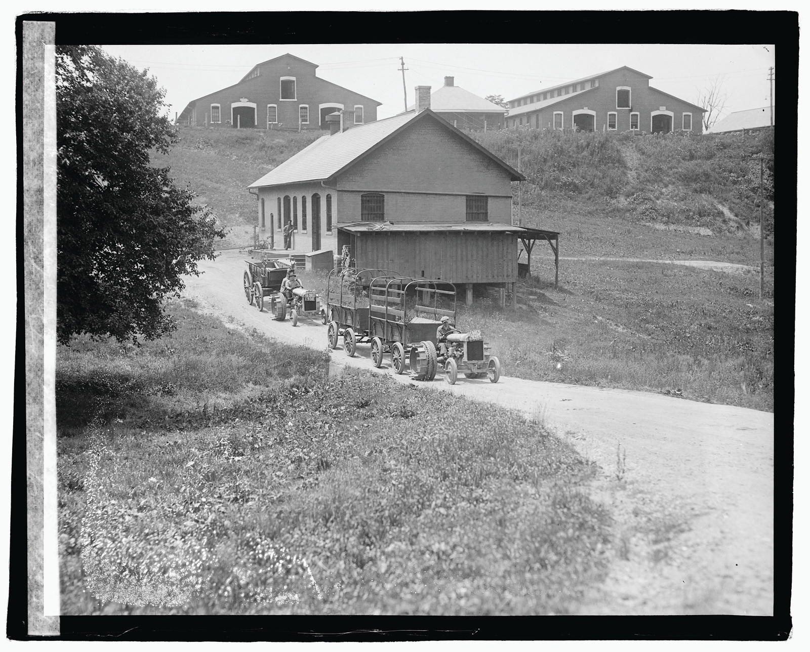 Two tractors pulling wagons on a farm road, barns behind — historic image