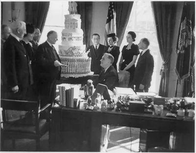 [Franklin Delano Roosevelt, seated by his White House desk surrounded by birthday cake and his staff]