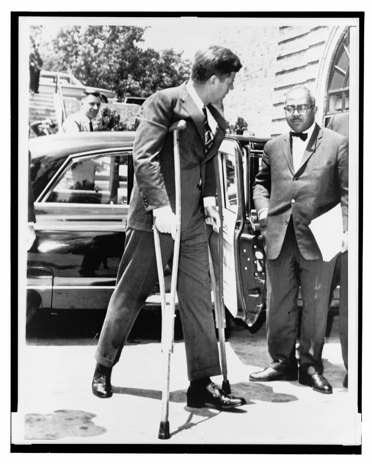 [President John F. Kennedy on crutches accompanied by White House Press Secretary Andrew T. Hatcher on way to a conference in Washington, D.C.], 1961 June 16. — historic photograph