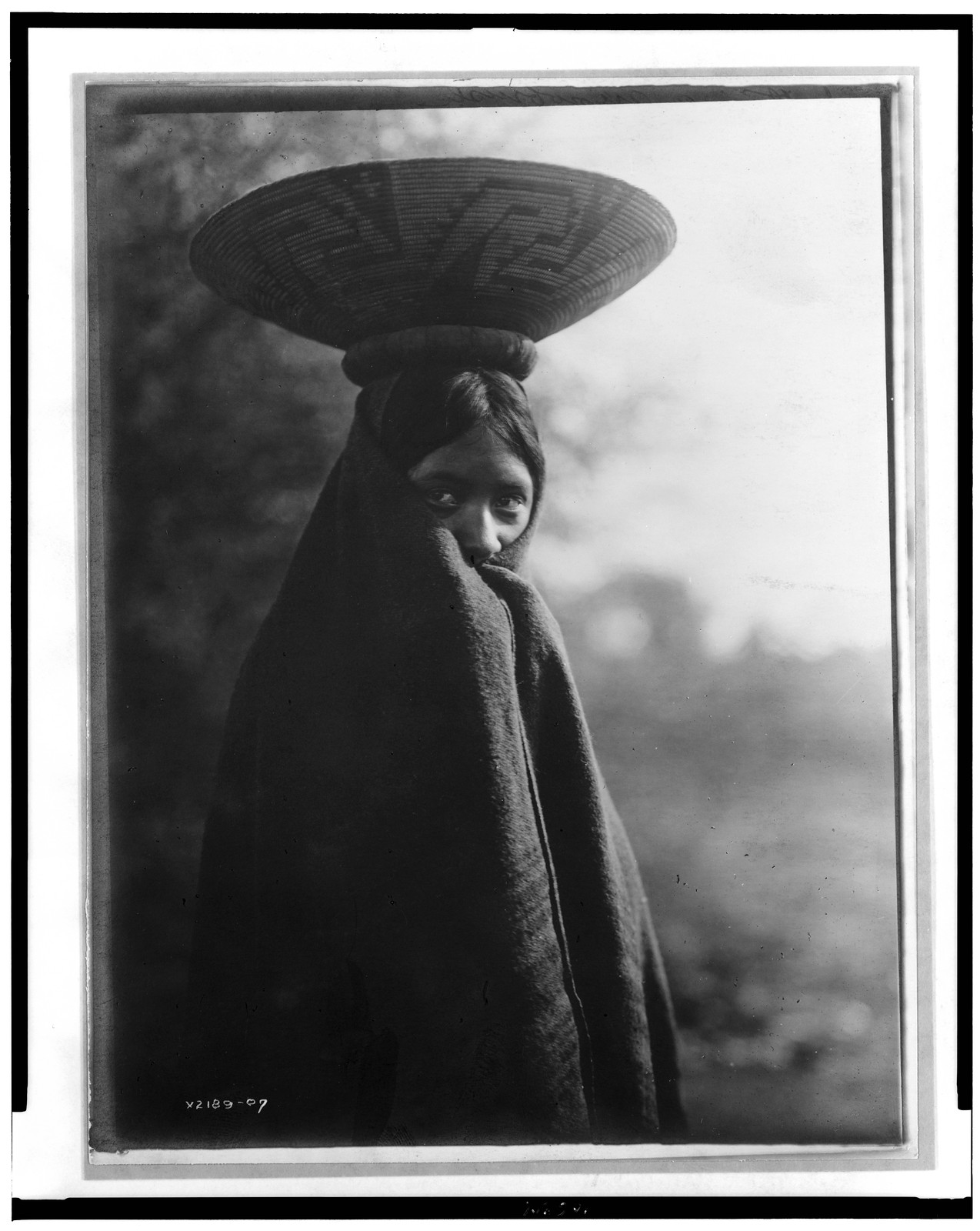 [Maricopa girl, half-length portrait, standing, facing slightly right, wrapped in blanket, with basket tray on head], c1907. — historic photograph