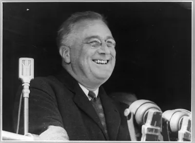 [Franklin Delano Roosevelt, head-and-shoulders portrait, facing right, at microphones, smiling]